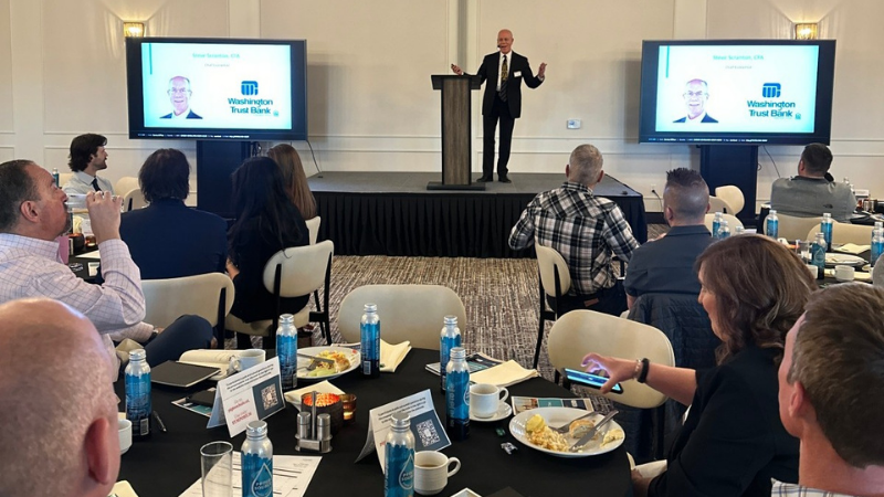 A speaker presenting in front of an audience at a conference, with food on tables, during the 2025 Financial Outlook & Tax Symposium recap.