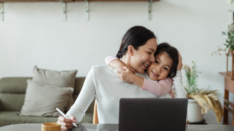 A woman and a girl smiling while the girl hugs the woman at a desk with a laptop.