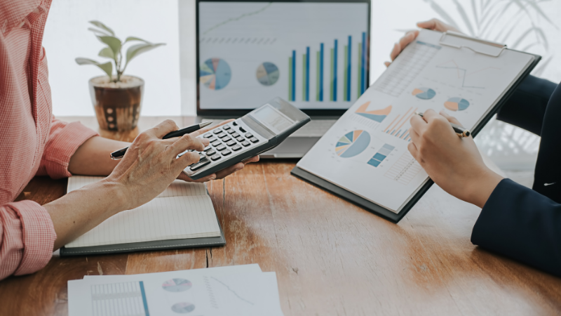 Two people at a wooden table discussing charts, data, and excess business losses, one using a calculator.