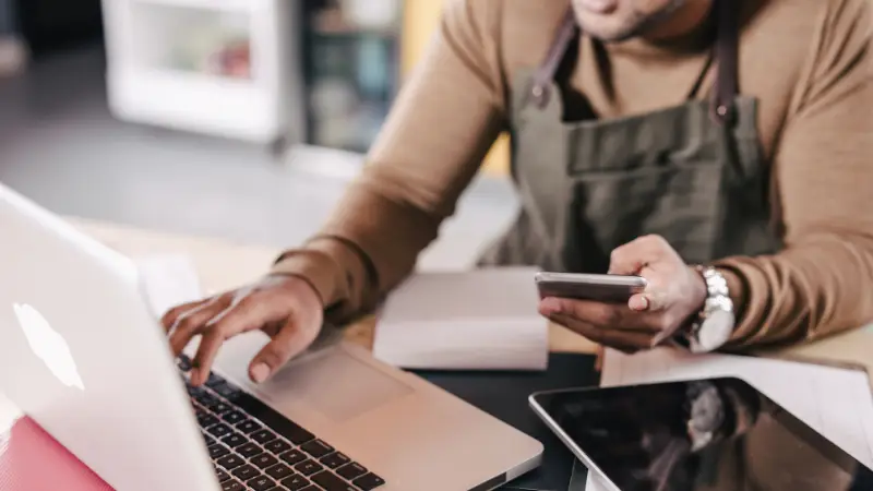 A person in a brown sweater and apron typing on a laptop while holding a smartphone, in need of an auditor to verify account balances and transactions.