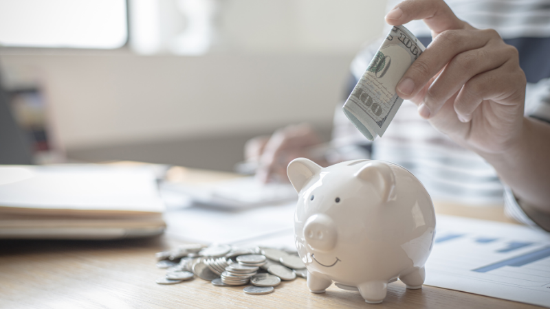 A person adds a rolled-up $100 bill into a white piggy bank on a desk with coins, highlighting how inflation impacts your taxes and returns.
