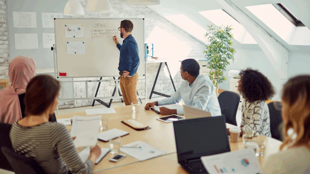 A group of people in a meeting room watching a man writing on a whiteboard and preparing for audit.
