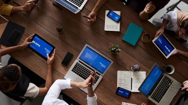 A group of people sit around a large wooden table engaged in a collaborative meeting. Laptops, tablets, notebooks, and coffee cups are spread across the table, with screens displaying charts and presentation slides. The setting suggests a modern, tech-focused business environment centered on teamwork and digital productivity.