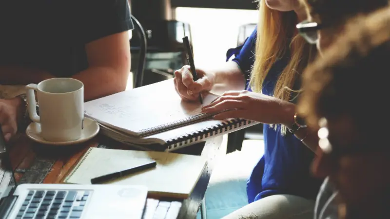 A group of people are gathered around a wooden table in a casual workspace. One person is writing in a spiral notebook, while others engage in discussion. The table holds a coffee mug, open notebooks, and a laptop, indicating a collaborative brainstorming or planning session. Natural light filters into the room, creating a warm and focused atmosphere.
