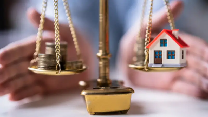 A close-up image of a brass balance scale shows coins stacked on one side and a small model house on the other, symbolizing financial decisions or weighing monetary value against property investment. A person's hands are positioned in the background, suggesting protection or deliberation. The scene conveys themes of real estate, asset management, or financial planning.
