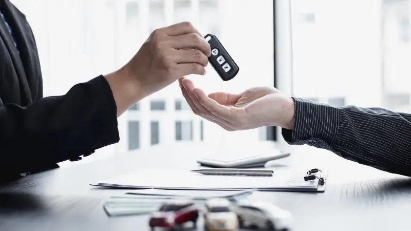 A person in business attire hands over a car key fob to another individual across a desk, symbolizing a car sale or lease agreement. The desk holds documents, a calculator, and small toy cars, suggesting a formal automotive transaction in a dealership or financial setting. The bright office environment adds a professional tone.
