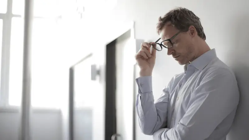 A man in a light blue dress shirt is leaning against a white wall in a brightly lit hallway. He appears deep in thought or stressed, with his head slightly bowed and his eyes closed, holding his glasses in one hand near his forehead.