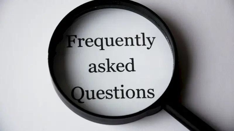 A magnifying glass hovers over the printed words "Frequently asked Questions" on a white sheet of paper, emphasizing the text. The image symbolizes searching for or highlighting important information, commonly associated with FAQ sections or informational resources.