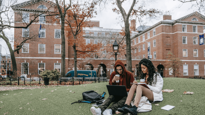 Two students studying on green grass under trees, with a brick building behind.
