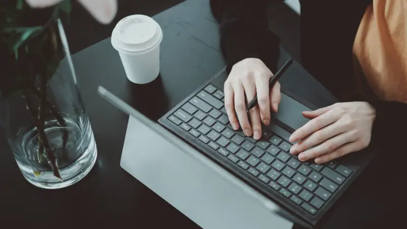 A person is working on a tablet with a detachable keyboard, using a stylus while typing. The workspace includes a black desk, a white coffee cup, and a vase with pink flowers, creating a clean and focused environment. The image conveys themes of remote work, digital productivity, or creative tasks.