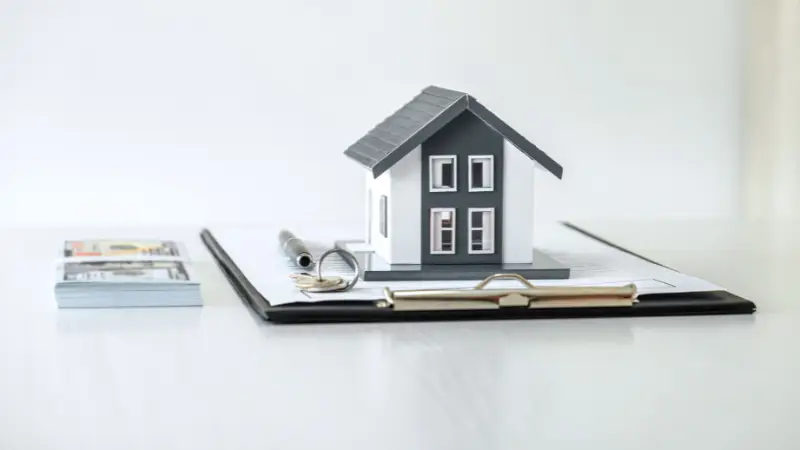 A small model house sits on a clipboard with a pen and keys, symbolizing real estate or home buying. Stacks of U.S. dollar bills are placed beside the clipboard on a white surface.