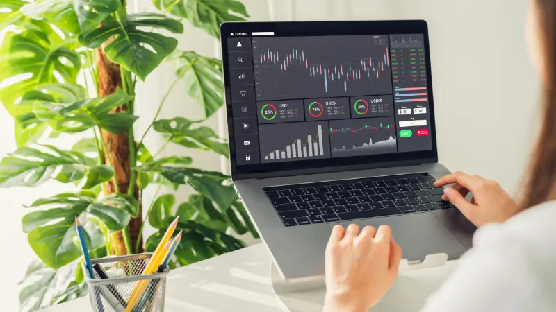 A person works on a laptop displaying a trading dashboard with charts and financial data. The setup is on a white desk with a green plant and a pencil holder containing colored pencils in the background.