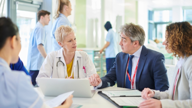 A medical team in a meeting with a doctor, a businessman, and a woman, discussing hospital accounting with a laptop and documents.