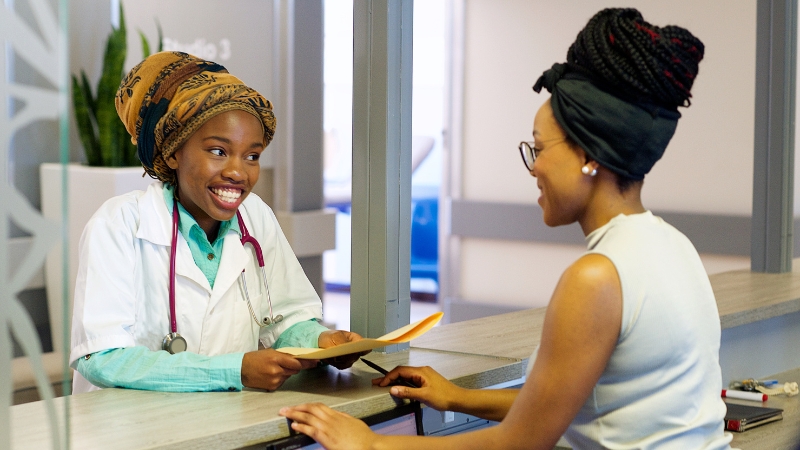 A smiling healthcare worker in a white coat discusses medical accounting to a woman at a reception desk.