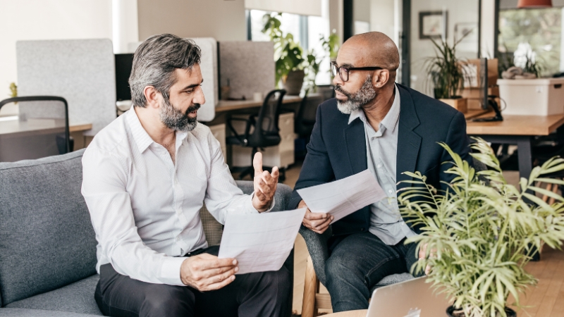 Two men discussing private wealth management documents while sitting on a couch in a modern office.