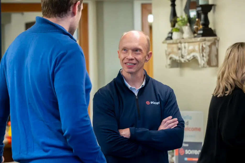 Two men talking in a room; one man smiles while the other listens, people in the background.